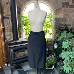 May include: A black pencil skirt with a belt, displayed on a mannequin. The skirt is knee-length and has a fitted silhouette. The mannequin is set against a backdrop of a stone wall and greenery.