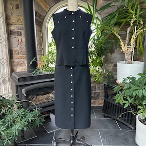 May include: A vintage black two-piece dress set. The sleeveless top has a small collar and white buttons. The matching skirt is knee-length with a row of white buttons down the front. The dress is displayed on a mannequin.