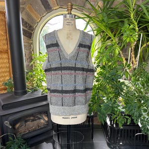 May include: A gray, blue, and red plaid V-neck sweater vest displayed on a mannequin. The vest has a ribbed bottom and a classic pattern. The mannequin is on a black metal stand, with a fireplace and plants in the background.