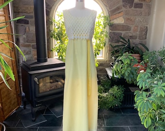 May include: A vintage yellow maxi dress on a mannequin. The bodice is white crocheted lace, and the skirt is a pale yellow with a subtle polka dot pattern. A matching yellow sash hangs down the front. The dress is displayed in front of a stone wall.