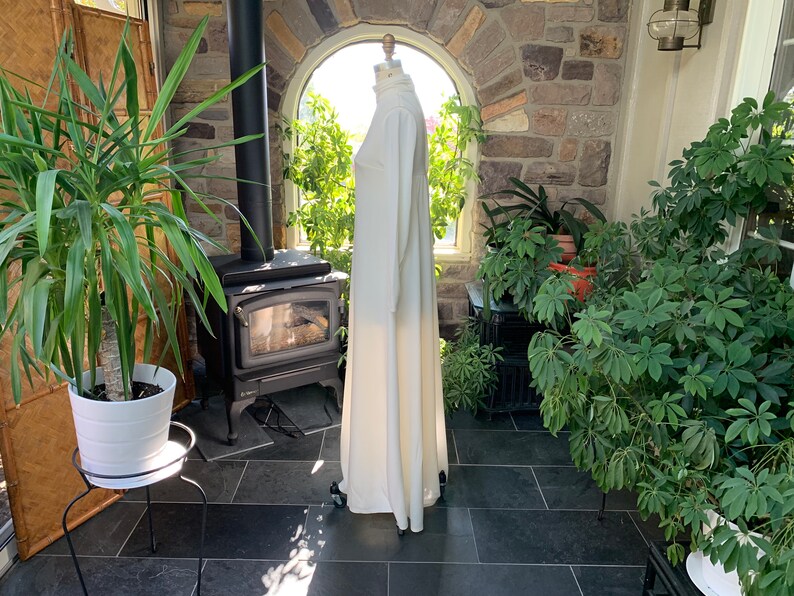 Peut inclure: Une longue robe blanc cass&eacute;, avec un col montant et des manches longues, est pr&eacute;sent&eacute;e sur un mannequin. La robe est longue et a une silhouette fluide. L'arri&egrave;re-plan comprend un po&ecirc;le noir, des plantes et un mur en pierre.