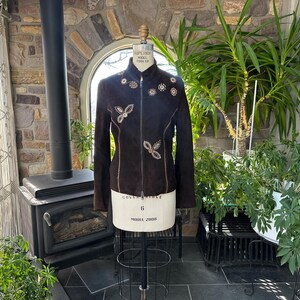 May include: A dark brown suede jacket with intricate floral and butterfly-shaped embellishments. The jacket features a high collar and a zipper closure. The design includes decorative stitching and is displayed on a mannequin. The jacket is a size 6.