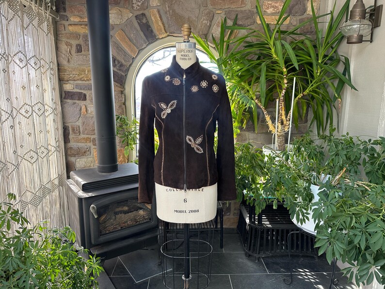 May include: A dark brown suede jacket with intricate floral and butterfly-shaped embellishments. The jacket features a high collar and a zipper closure. The design includes decorative stitching and is displayed on a mannequin. The jacket is a size 6.