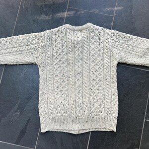 K&ouml;nnte beinhalten: Ein hellgrauer Zopfstrickpullover mit Rundhalsausschnitt. Der Pullover hat aufwendige Webmuster und gesprenkelte Details. Die langen &Auml;rmel und das klassische Design machen ihn zu einem warmen Kleidungsst&uuml;ck. Ideal f&uuml;r k&uuml;hlere Tage.