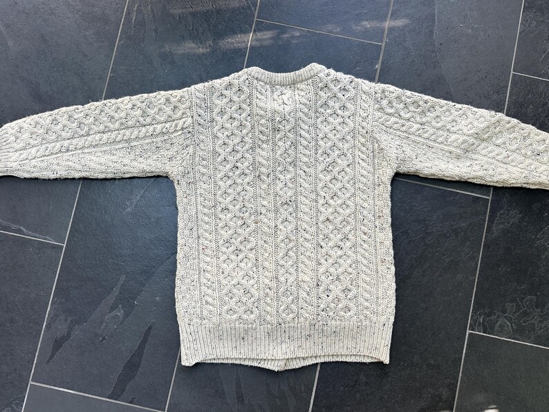 K&ouml;nnte beinhalten: Ein hellgrauer Zopfstrickpullover mit Rundhalsausschnitt. Der Pullover hat aufwendige Webmuster und gesprenkelte Details. Die langen &Auml;rmel und das klassische Design machen ihn zu einem warmen Kleidungsst&uuml;ck. Ideal f&uuml;r k&uuml;hlere Tage.