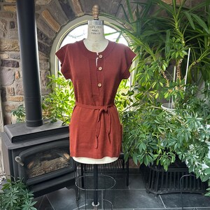 May include: A rust-colored knit dress with short sleeves and a matching belt. The dress features a button-down front and is displayed on a mannequin. The background includes a fireplace, plants, and a window.