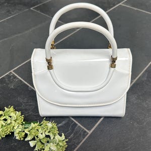 May include: A white patent leather handbag with a glossy finish, featuring a structured rectangular body, a curved flap closure, and two rounded top handles attached with gold-toned metal hardware rings at the base of each handle.