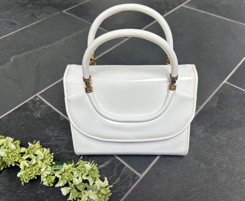 May include: A white patent leather handbag with a glossy finish, featuring a structured rectangular body, a curved flap closure, and two rounded top handles attached with gold-toned metal hardware rings at the base of each handle.