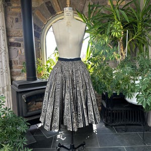 May include: A vintage black and white patterned skirt displayed on a mannequin. The skirt has a pleated design with a repeating geometric pattern. The skirt is displayed in front of a window with plants.