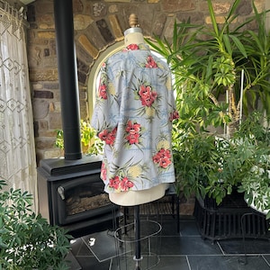 Może przedstawiać: Jasnoniebieska koszula hawajska z kwiatowym i ananasowym wzorem. Koszula ma kr&oacute;tkie rękawy i zapięcie na guziki z przodu. Wz&oacute;r zawiera czerwone kwiaty i ż&oacute;łte ananasy.