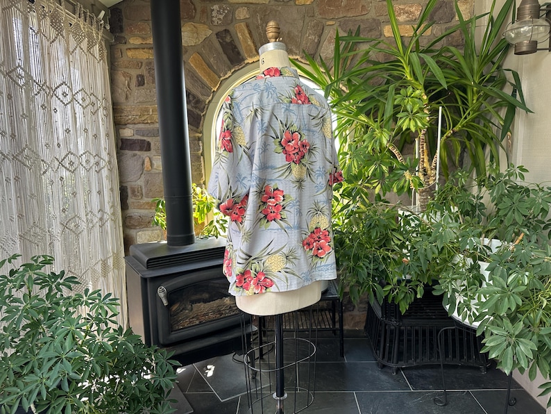 Może przedstawiać: Jasnoniebieska koszula hawajska z kwiatowym i ananasowym wzorem. Koszula ma kr&oacute;tkie rękawy i zapięcie na guziki z przodu. Wz&oacute;r zawiera czerwone kwiaty i ż&oacute;łte ananasy.