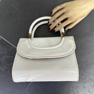 May include: A glossy white patent leather handbag featuring a structured rectangular body, a curved flap closure, and a single rounded handle attached with gold-toned metal hardware. The surface has a reflective finish and clean lines.