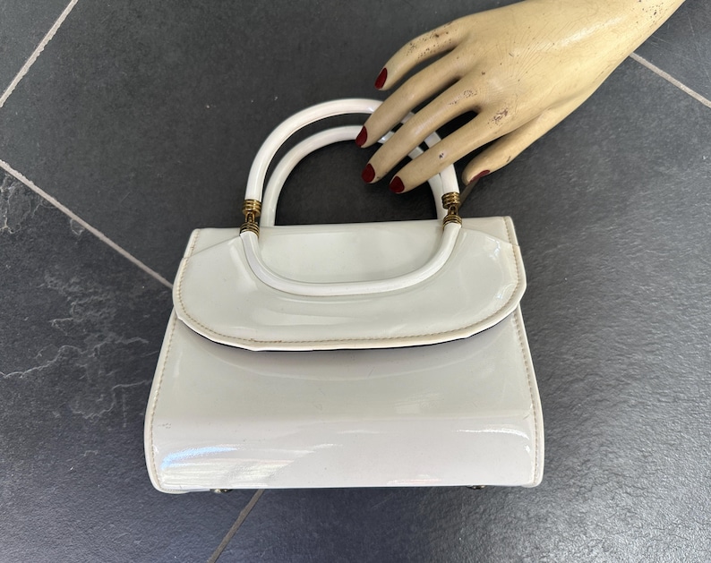 May include: A glossy white patent leather handbag featuring a structured rectangular body, a curved flap closure, and a single rounded handle attached with gold-toned metal hardware. The surface has a reflective finish and clean lines.