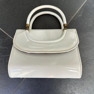 May include: A glossy white handbag with a structured rectangular body and a curved flap closure. It features two rounded top handles attached with metallic gold-toned hardware accents at the base of each handle, providing a polished finish.