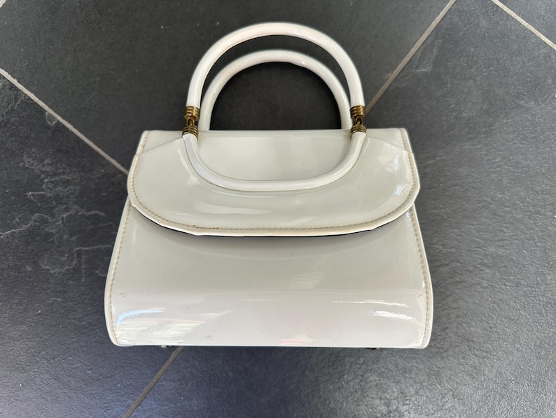 May include: A glossy white handbag with a structured rectangular body and a curved flap closure. It features two rounded top handles attached with metallic gold-toned hardware accents at the base of each handle, providing a polished finish.