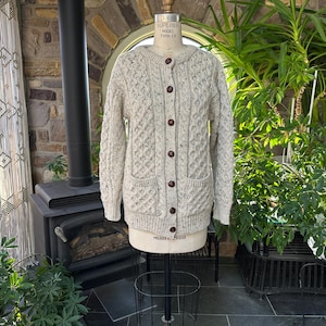 K&ouml;nnte beinhalten: Hellbeige Cardigan-Strickjacke mit Knopfleiste und Zopfmuster. Der Pullover hat zwei Vordertaschen und braune Kn&ouml;pfe. Der Pullover wird auf einer Schaufensterpuppe pr&auml;sentiert. Der Pullover ist Gr&ouml;&szlig;e Medium.