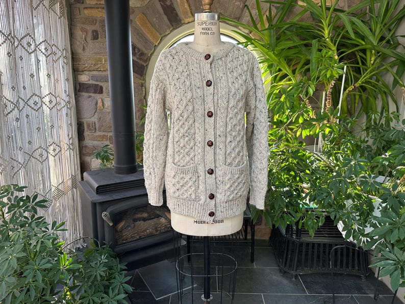 K&ouml;nnte beinhalten: Hellbeige Cardigan-Strickjacke mit Knopfleiste und Zopfmuster. Der Pullover hat zwei Vordertaschen und braune Kn&ouml;pfe. Der Pullover wird auf einer Schaufensterpuppe pr&auml;sentiert. Der Pullover ist Gr&ouml;&szlig;e Medium.