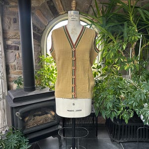 Puede incluir: Un chaleco beige con botones y textura acanalada, acentuado con ribetes rojos, verdes y negros a lo largo del escote y las sisas. El chaleco se exhibe en un maniquí, mostrando su estilo vintage. Tiene cuello en V y es sin mangas.