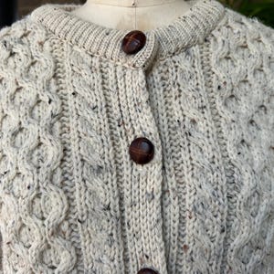 K&ouml;nnte beinhalten: Cremefarbene Strickjacke mit Zopfmuster und braunen Kn&ouml;pfen. Der Pullover hat einen runden Ausschnitt und ein strukturiertes, gemustertes Design. Die Kn&ouml;pfe sind dunkelbraun. Der Pullover ist aus dickem, warmem Material.