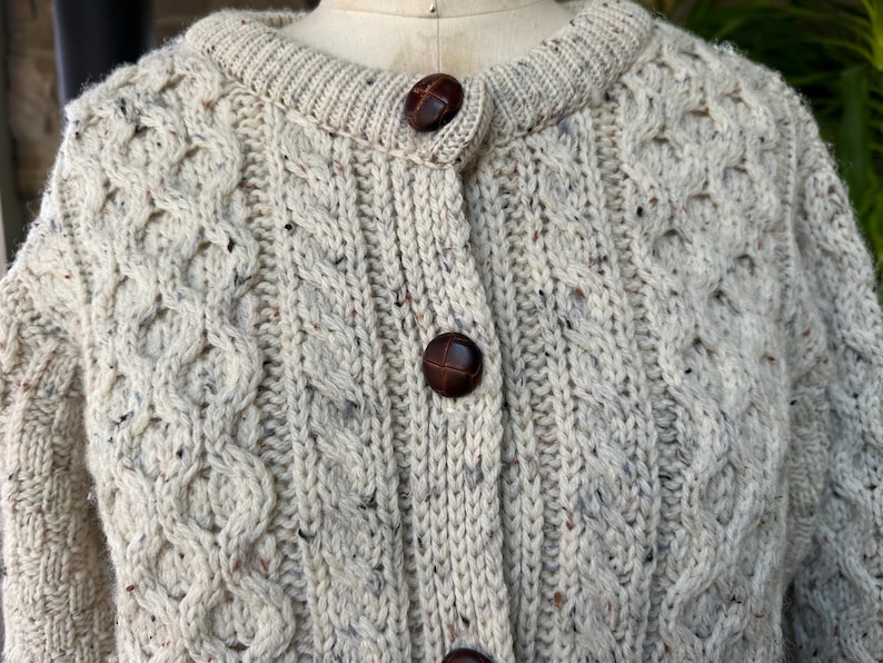 K&ouml;nnte beinhalten: Cremefarbene Strickjacke mit Zopfmuster und braunen Kn&ouml;pfen. Der Pullover hat einen runden Ausschnitt und ein strukturiertes, gemustertes Design. Die Kn&ouml;pfe sind dunkelbraun. Der Pullover ist aus dickem, warmem Material.