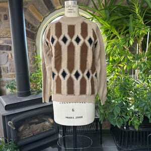 May include: A beige sweater with a geometric pattern of brown vertical stripes and black diamond shapes. The sweater is displayed on a mannequin. The sweater has a ribbed neckline, cuffs, and hem. The mannequin is on a black metal stand.