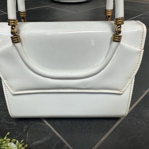 May include: A glossy white patent leather handbag featuring a structured rectangular body with a flap closure. The bag includes two white arched handles attached to the top with gold-toned metal hardware rings and ribbed cylindrical accents.
