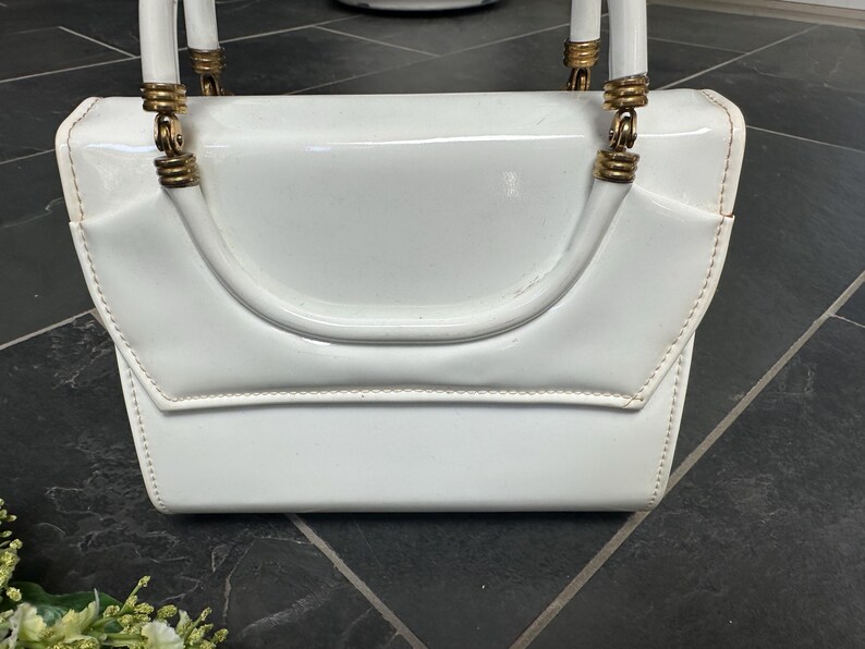 May include: A glossy white patent leather handbag featuring a structured rectangular body with a flap closure. The bag includes two white arched handles attached to the top with gold-toned metal hardware rings and ribbed cylindrical accents.