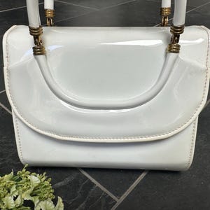 May include: A white patent leather handbag with a glossy finish, featuring a structured flap closure and a single top handle. The handle is attached to the bag with gold-toned metal hardware rings, providing a polished contrast to the white surface.