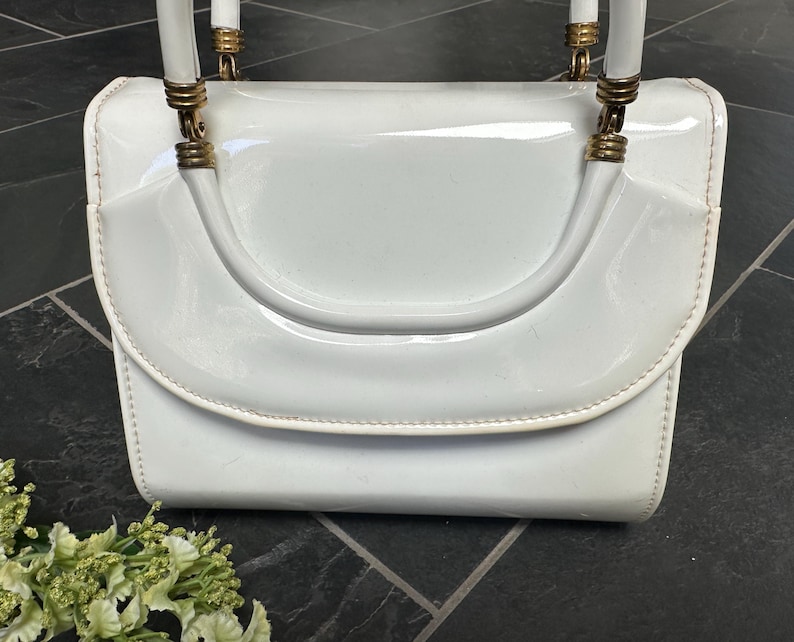 May include: A white patent leather handbag with a glossy finish, featuring a structured flap closure and a single top handle. The handle is attached to the bag with gold-toned metal hardware rings, providing a polished contrast to the white surface.