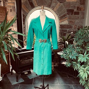 Puede incluir: Un abrigo largo de color turquesa con cuello con solapas y cierre de botones. El abrigo presenta un cinturón a juego con una hebilla decorativa. El abrigo se exhibe en un maniquí. La longitud del abrigo es de aproximadamente 114 cm.