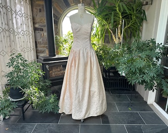 Könnte beinhalten: Ein vintage, trägerloses, hellpfirsichfarbenes Kleid auf einer Schaufensterpuppe. Das Kleid hat ein eng anliegendes Oberteil und einen weiten Rock mit floralem Muster. Das Kleid wird in einer sonnigen Umgebung im Freien mit Grün und einem Steinkamin im Hintergrund präsentiert.