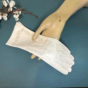 Könnte beinhalten: Ein Paar weiße Lederhandschuhe mit zarter Blumenstickerei. Die Handschuhe sind in der Mitte gefaltet und werden von einer Handpuppe gehalten.