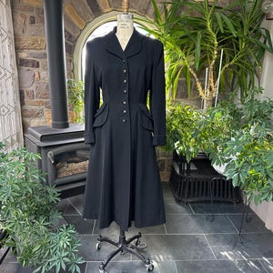 May include: A long, black coat with a tailored fit, displayed on a dress form. The coat has a button-down front, a notched collar, and pockets. The coat is a classic style, suitable for various occasions.