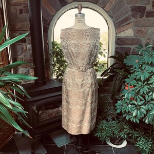 Peut inclure: Robe beige vintage à motif géométrique, présentée sur un mannequin. La robe sans manches présente une silhouette ajustée et une ceinture assortie avec une boucle rectangulaire. La robe est en tissu texturé, sur fond de plantes et de mur en pierre.