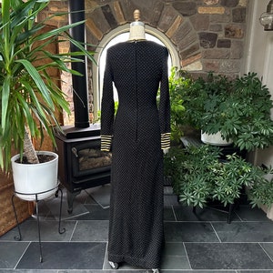 Pu&ograve; includere: Un abito nero lungo fino al pavimento con piccoli pois bianchi. Le maniche lunghe e lo scollo sono rifiniti con strisce dorate. L'abito &egrave; esposto su un manichino, evidenziando il suo design elegante. L'abito &egrave; in un ambiente esterno ben illuminato.