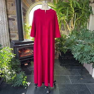 Könnte beinhalten: Ein langes, leuchtend rotes Kleid mit einem halblangen Reißverschluss und Dreiviertelärmeln wird auf einer Schaufensterpuppe präsentiert. Das Kleid ist bodenlang und aus einem einfarbigen, glatten Stoff gefertigt. Die Schaufensterpuppe steht auf Rollen, und der Hintergrund umfasst einen Kamin und Grünpflanzen.