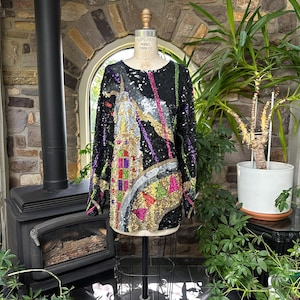 May include: A long-sleeved black top covered in multicoloured sequins featuring a prominent architectural design. The garment displays a tall, silver-toned structure with accents of gold, pink, purple, and green sequins arranged in geometric patterns.