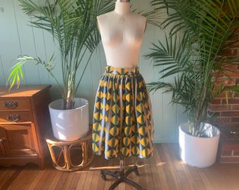 May include: A patterned skirt displayed on a mannequin. The skirt features a repeating geometric design in yellow, teal, and gray. The skirt is knee-length and has a gathered waist. The mannequin is set against a backdrop of plants and a wooden floor.