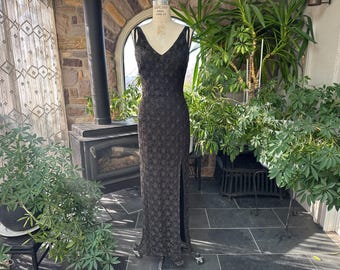May include: A floor-length, black evening gown with a V-neck and spaghetti straps. The dress features a subtle, all-over textured pattern and a high slit on the left side. The dress is displayed on a mannequin.