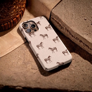 May include: A phone case with a repeating pattern of brown horses on a light beige background. The case has a camera cutout with three lenses and a flash. The case is lying on a brown surface.