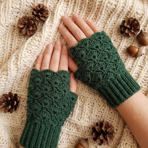 Könnte beinhalten: Ein Paar dunkelgrüne Fingerhandschuhe mit gewelltem Rand und einem strukturierten Häkelmuster. Die Handschuhe werden auf einer cremefarbenen Strickoberfläche mit Kiefernzapfen und Eicheln präsentiert.