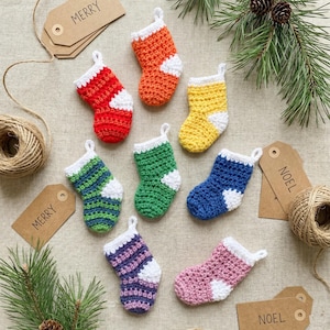 Könnte beinhalten: Eine Sammlung bunter, gehäkelter Miniatur-Weihnachtssocken in verschiedenen Farben, darunter Rot, Orange, Gelb, Grün, Blau, Lila und Rosa. Jede Socke hat eine weiße Manschette und Ferse. Braune Etiketten mit "MERRY" und "NOEL" sind zu sehen.
