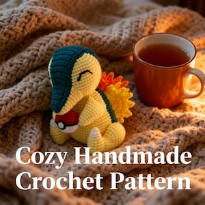 Può includere: Un peluche Cyndaquil all'uncinetto fatto a mano in giallo, verde e arancione, che tiene una Poké Ball rossa e bianca. Il peluche è su una coperta accogliente e strutturata accanto a una tazza di tè arancione. Testo: "Cozy Handmade Crochet Pattern."