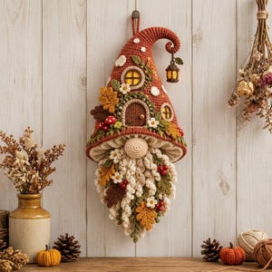 May include: A crocheted wall hanging shaped like a gnome with a tall, pointed rust-coloured hat featuring a small door, windows, and a hanging lantern. The gnome has a cream-coloured beard decorated with crocheted autumn leaves, white flowers, and red berries.