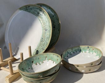 May include: A set of handmade ceramic tableware, including plates and bowls. The plates have a white center with a seafoam green rim. The bowls are seafoam green and white. The pottery has a speckled design and a rustic aesthetic.