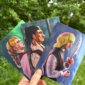 Könnte beinhalten: Drei rechteckige Kunstdrucke mit stilisierten digitalen Illustrationen von Charakteren. Der linke Druck zeigt eine Person mit blondem Zopf, der mittlere Druck eine Person mit dunklem Pferdeschwanz und der rechte Druck eine blonde Person.