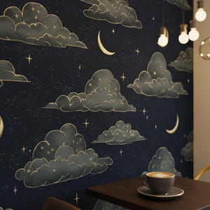 Op de afbeelding: Donkerblauwe behang met een hemels ontwerp van goudomrande wolken, halve manen en sterren. Het herhalende patroon creëert een dromerige nachthemel-esthetiek. Een kopje koffie staat op een donkerbruine tafel.