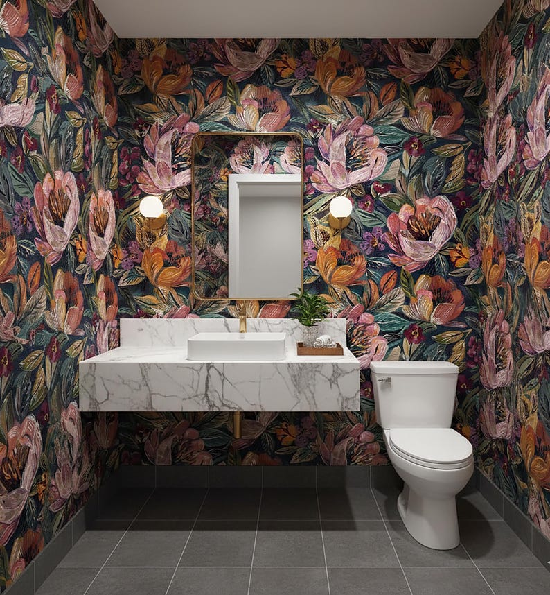 May include: A bathroom with floral wallpaper in shades of pink, orange, and green. The room features a white marble vanity with a rectangular sink, a gold-framed mirror, and a white toilet. Two wall lights flank the mirror.
