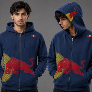Op de afbeelding: Een marineblauwe hoodie met rits, voorzien van een grote rode stierafbeelding op de voorzijde en een kleiner rood stierlogo op de borst. Het ontwerp bevat een geel cirkelvormig patroon achter de stier en een logo op de capuchon.