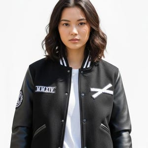 K&ouml;nnte beinhalten: Schwarze Collegejacke mit wei&szlig;en Akzenten, Leder&auml;rmeln und einem wei&szlig;en "X" Aufn&auml;her. Die Jacke hat einen wei&szlig;en Kragen mit schwarzen Streifen und einen Aufn&auml;her mit der Aufschrift "MMXIV".